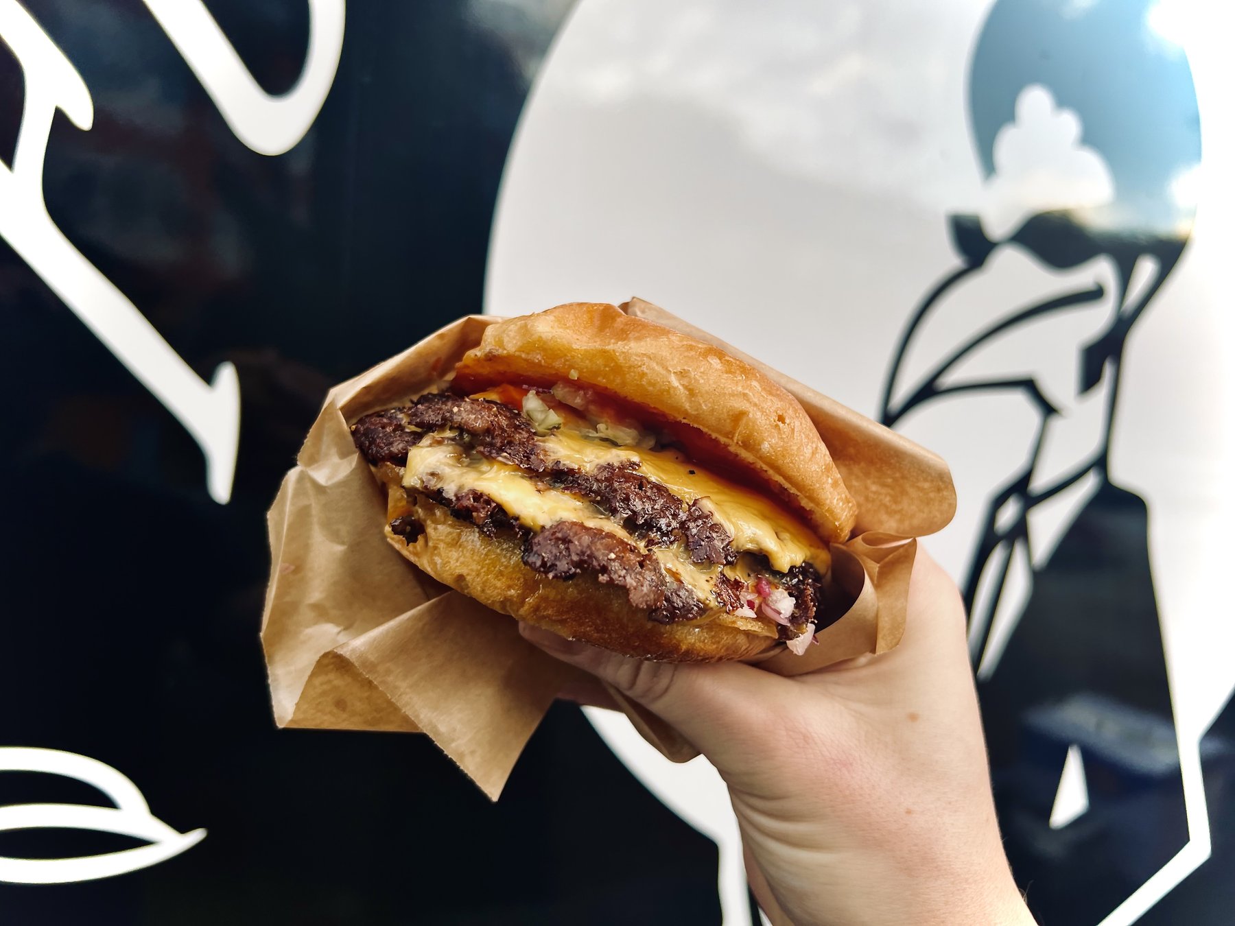 A hand holding a double smash cheeseburger wrapped in brown paper, in front of the Funky Chicken food truck