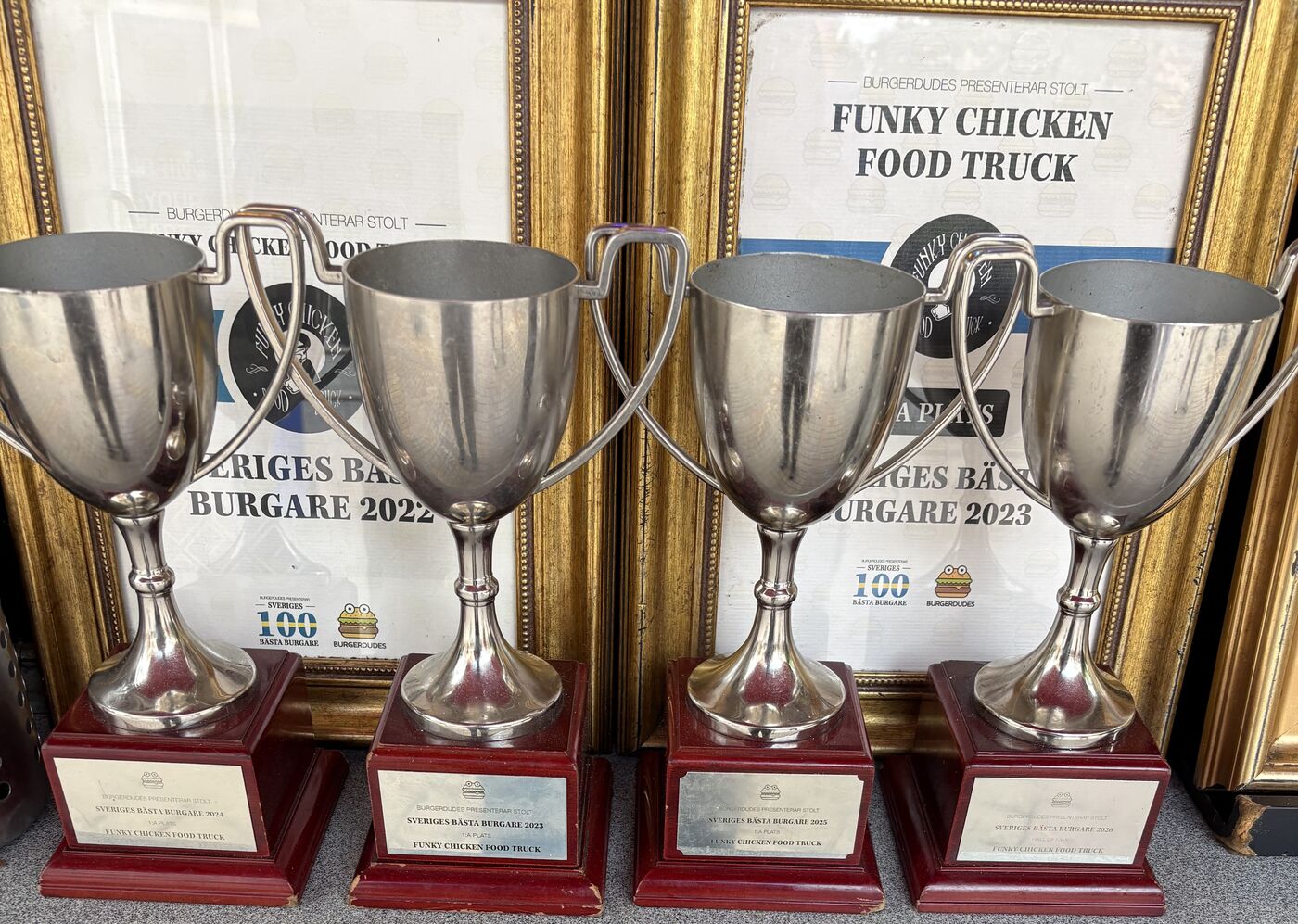 Four silver trophies on red wooden bases in front of framed certificates – Sveriges bästa burgare 1st place 2023, 2024 and 2025, and Hall of Fame 2026